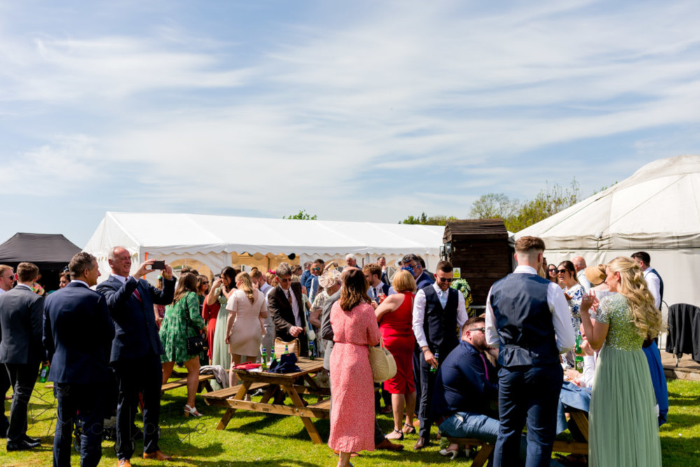 An image of a group of people smiling in the sunshine at a wedding at Salix Yurts, in the background marquees can be seen