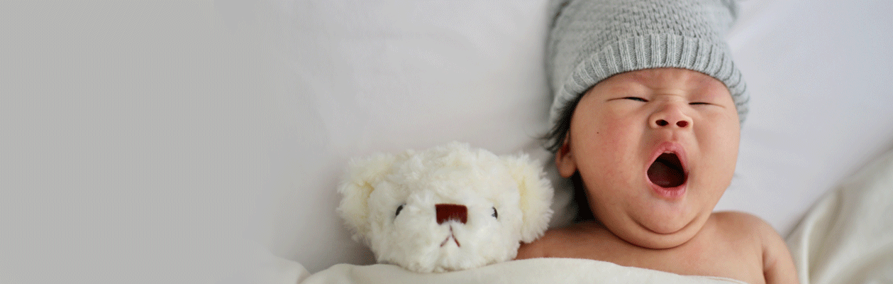 A baby in a grey hat yawning next to a white teddy bear