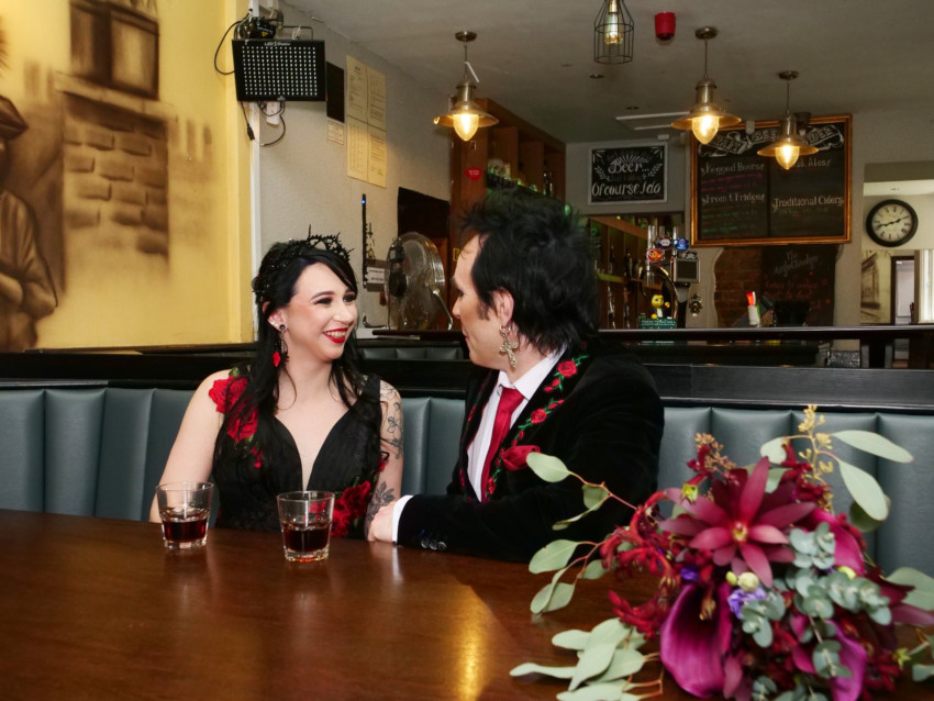 Bride and groom share a drink, they sit smiling together with the bouquet on the table