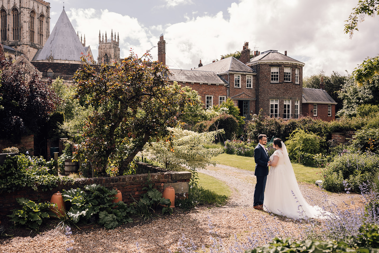 Exterior view of Grays Court from the gardens, a bride and groom stand facing each other in the centre of the garden, next to a large patch of vibrant flowers