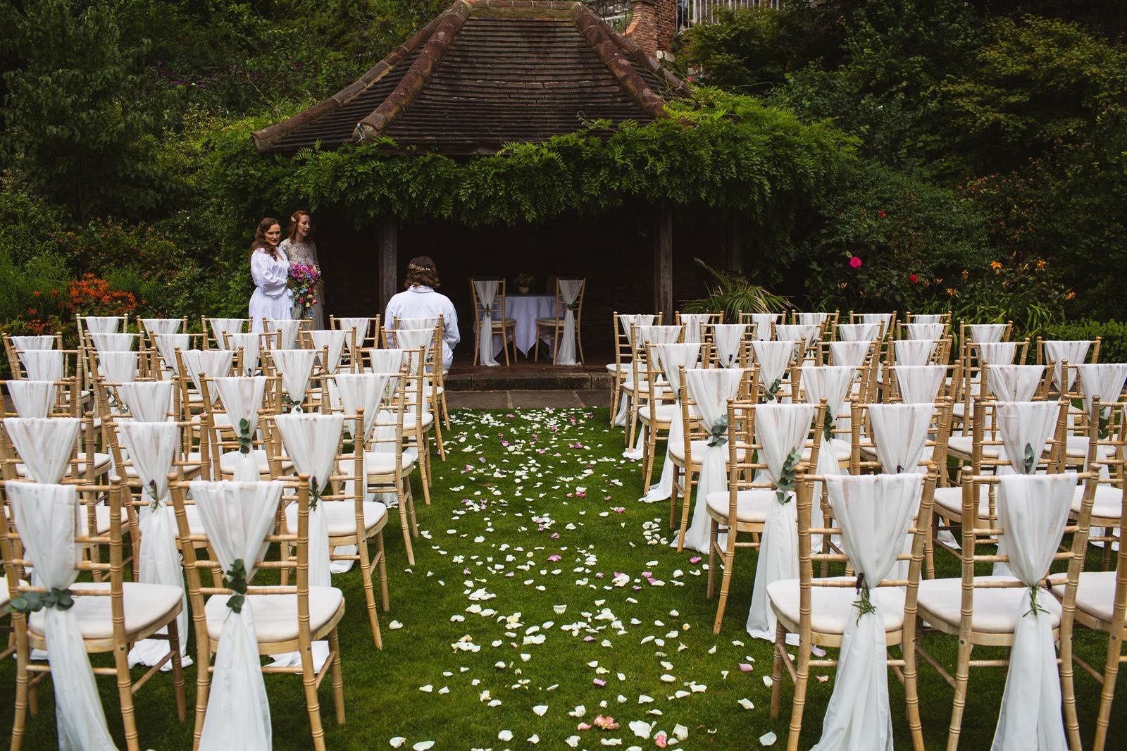 A view of chairs set up outside, facing a grandstand, with an aisle down the middle with petal spread on the floor