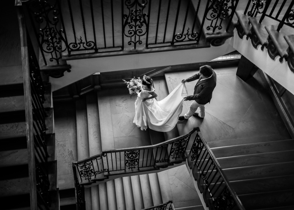 A black and white photograph of a bride walking down a staircase with a gentleman holding her train, the image is taken from above