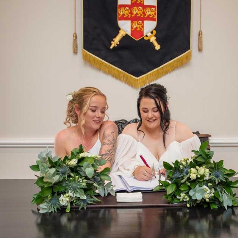 Two brides sat at a table which has their bouquets laid on are signing the marriage register. In the background the City of York Council logo can be seen